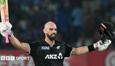 Daryl Mitchell holds his bat and helmet aloft after reaching his century against India
