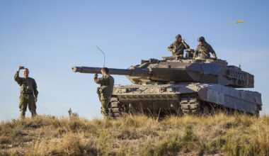 Soldiers stand and communicate beside a Leopard battle tank positioned on open terrain during daylight, with crew members visible on the turret and the main gun facing forward under a clear sky