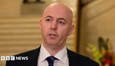 A bald man with short red stubble looks to the left of the frame. He wears a balck suit and purple and navy tie. He is standing in the Great Hall at Stormont in front of cream stairs, addressing the press