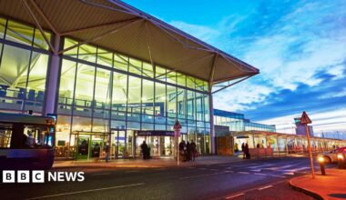 The exterior of the main terminal building at Bristol Airport. It is a large, modern structure with full-length glass windows, and the picture has been taken at dusk, with a dark blue, slightly cloudy sky. Passengers can be seen entering the terminal.