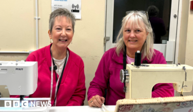 Two women sit with their sewing machines. The lady on the left has short grey hair and is wearing a bright pink jumper. She has dangly earrings and wears a tape measure around her neck. The lady on the left has longer grey hair with a fringe. A pair of glasses rests on her head. She is wearing a purple zip-up fleece. A floral sewing box is at the front of the image.