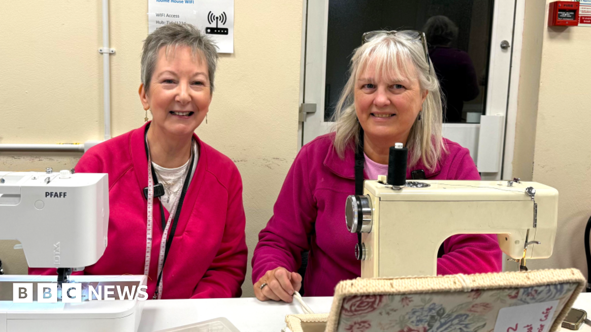 Two women sit with their sewing machines. The lady on the left has short grey hair and is wearing a bright pink jumper. She has dangly earrings and wears a tape measure around her neck. The lady on the left has longer grey hair with a fringe. A pair of glasses rests on her head. She is wearing a purple zip-up fleece. A floral sewing box is at the front of the image.