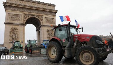Several tractors, including a red tractor flying French flags, are parked in front of the Arc de Triomphe in Paris.