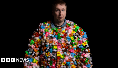Joe Lycett is wearing a costume made entirely of tine little stuffed toys that are multi-coloured. The animals include pink llamas, yellow ducks and blue hippos