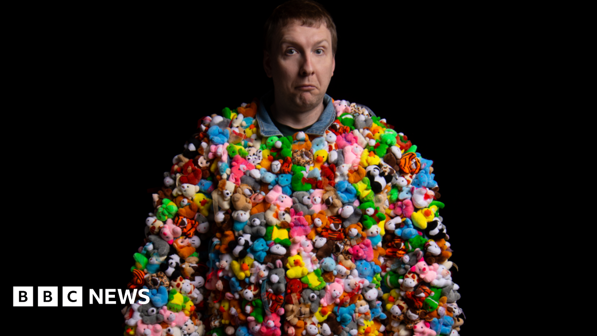 Joe Lycett is wearing a costume made entirely of tine little stuffed toys that are multi-coloured. The animals include pink llamas, yellow ducks and blue hippos