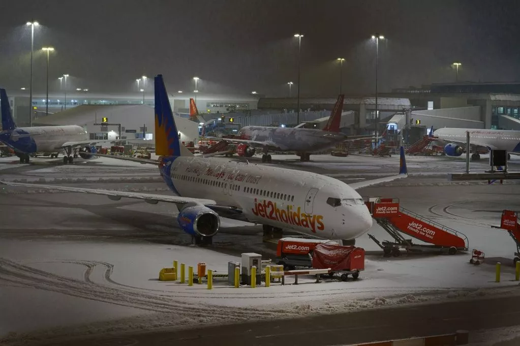 An airplane is parked on the runway at an airport during nighttime, with snow covering the ground and several airport vehicles nearby.