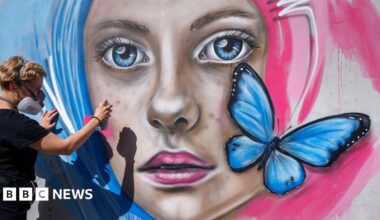 A woman paints a mural of a woman with pink and blue hair. The mural has a blue butterfly on it.