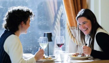 A queer couple has a meal at a fine dining restaurant. One person faces the camera and laughs at the other, who sits on the left away from camera smiling. On the table is food and wine. The person laughing holds a fork in hand and wears a white vest. The other has a black vest.