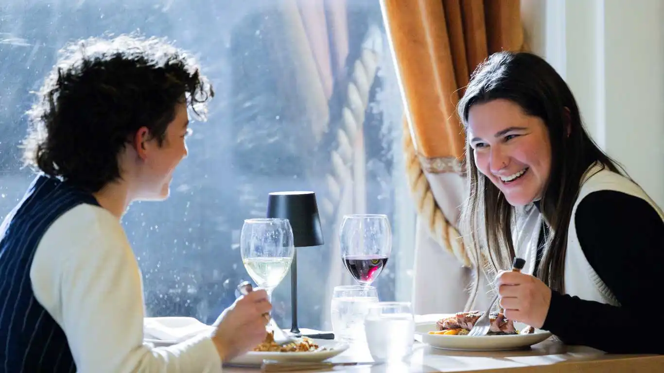 A queer couple has a meal at a fine dining restaurant. One person faces the camera and laughs at the other, who sits on the left away from camera smiling. On the table is food and wine. The person laughing holds a fork in hand and wears a white vest. The other has a black vest.