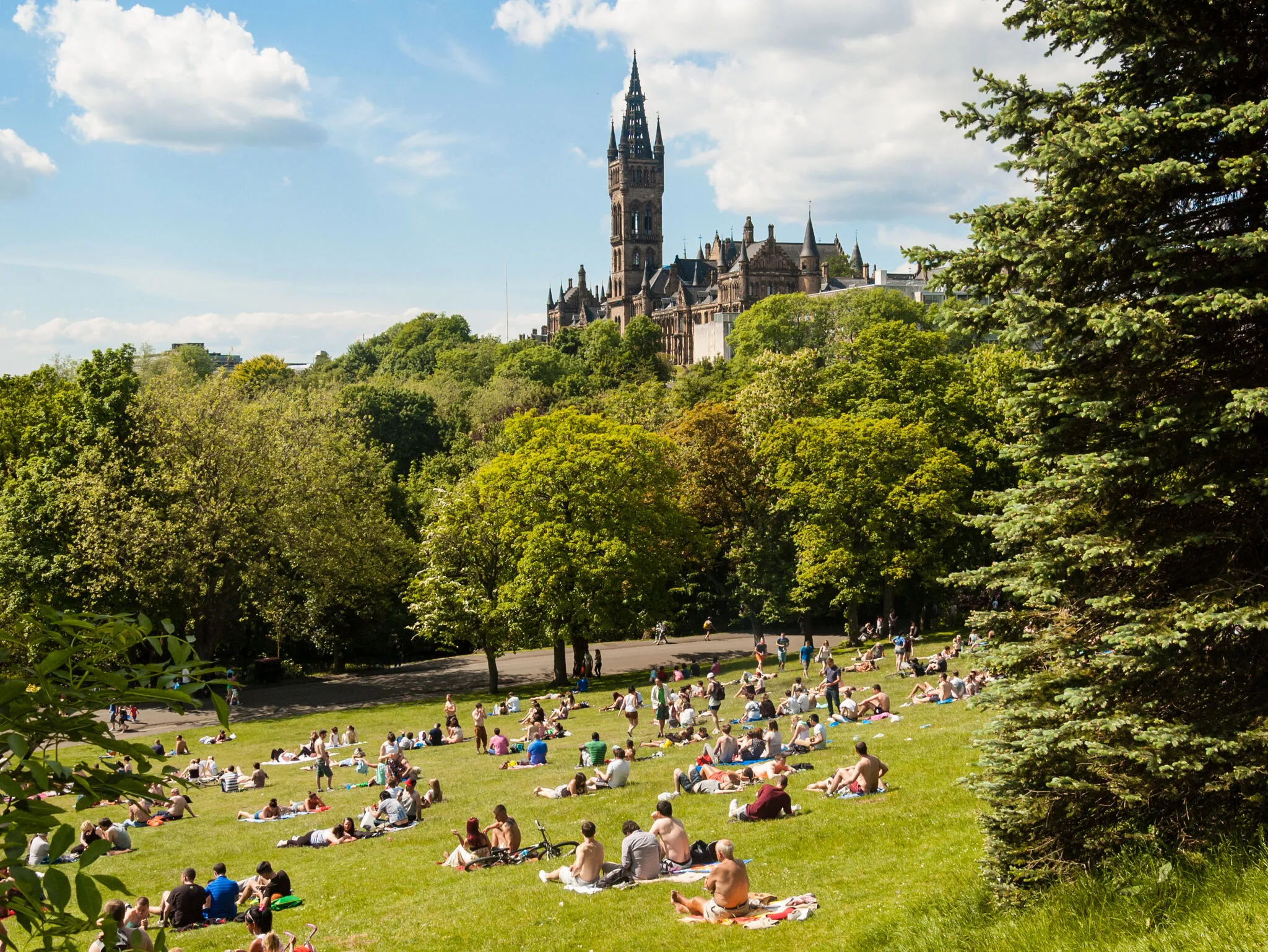 View of Kelvingrove Park full of people enjoying the Scottish summer with the main building of Glasgow University on the top of the hill.