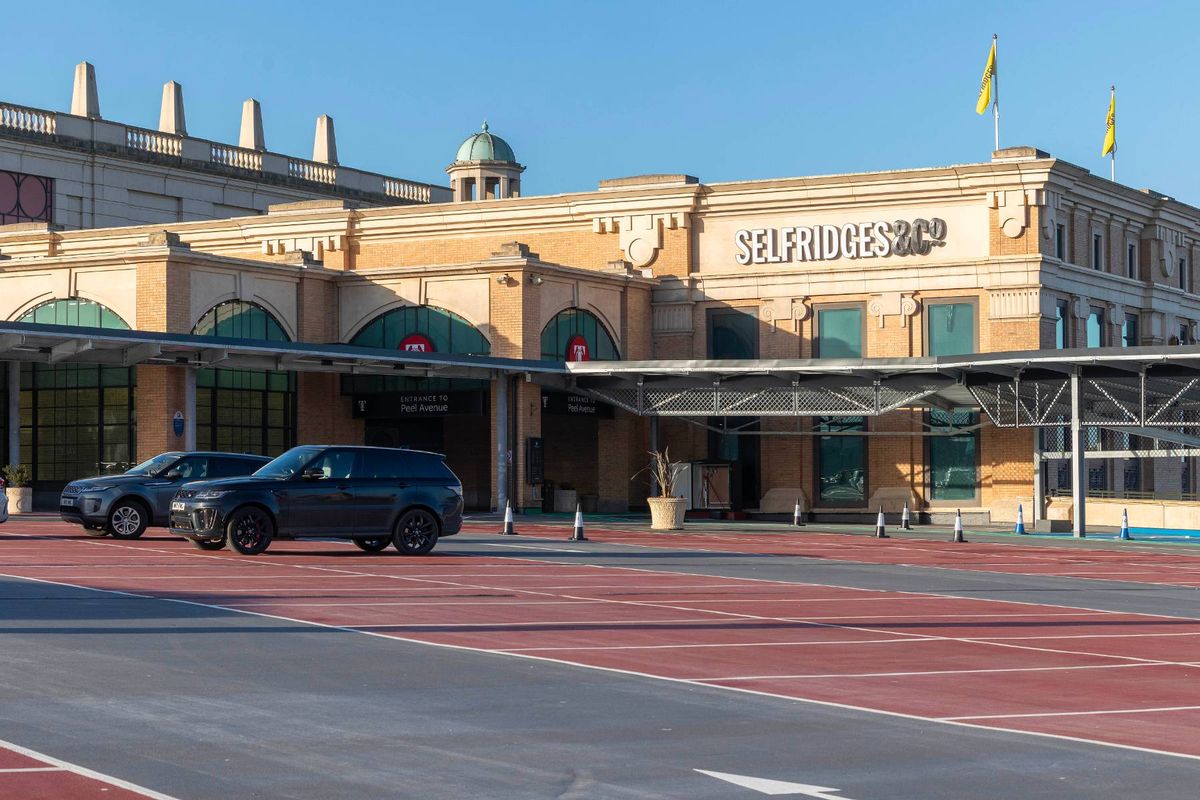Exterior view of a modern building with a large sign displaying the name "MARKS" in front of a parking area. Two cars are parked, and a person is visible near the entrance. The building has a classical architectural style with a dome roof and is situated against a clear blue sky.