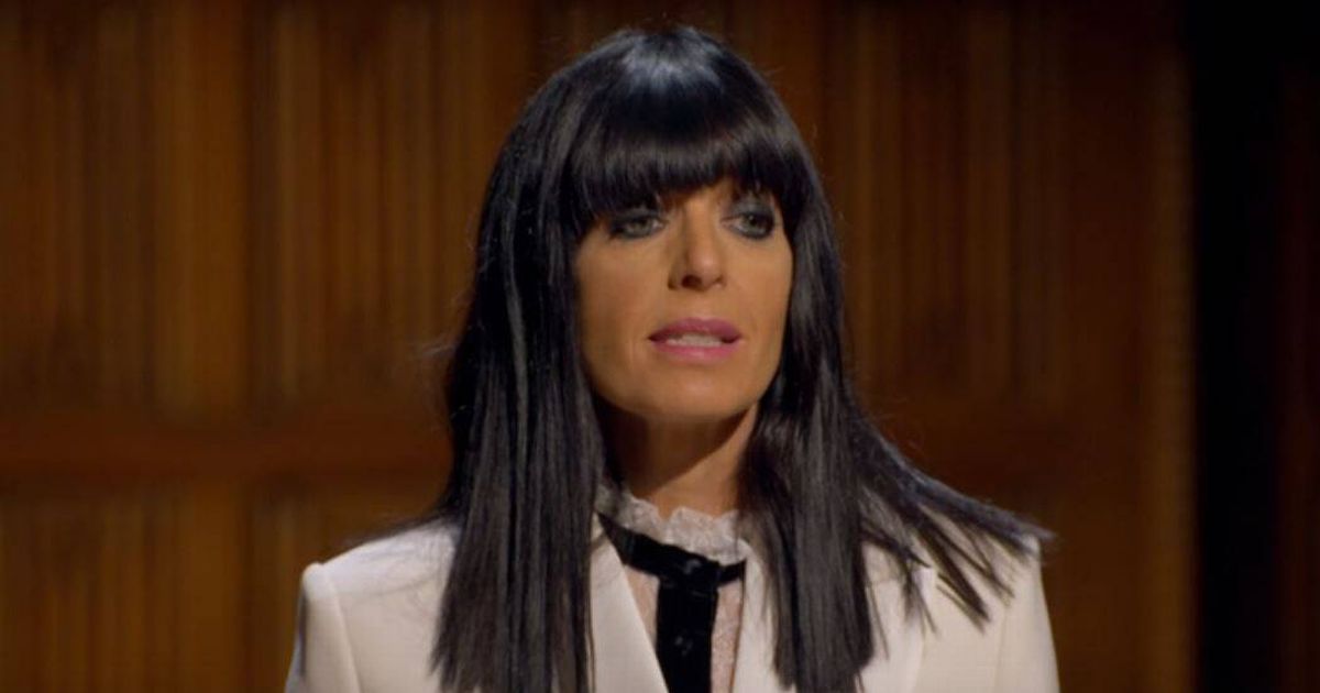 An individual with a beard, donning a green hooded cloak, stands in a dimly lit room with an intricate background.