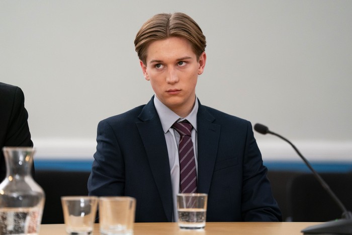 545911,TITLE:Eastenders Joel Marshall, in a blue suit and burgundy tie, sits in court with a microphone and some glasses of water in front of him in a scene from EastEnders
