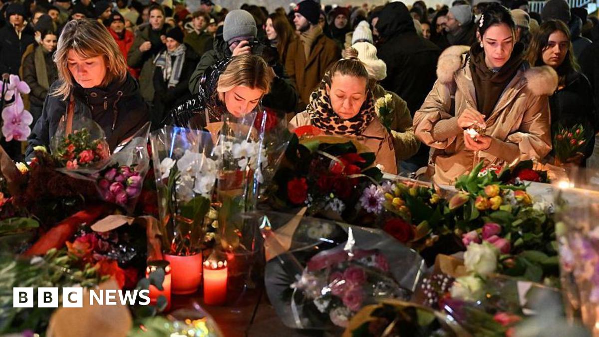People gather at the scene of a deadly bar fire in Switzerland. They are stood in front of a huge pile of flowers and candles.