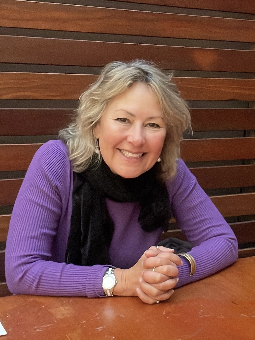 A middle aged white woman with blonde hair sitting in a courtyard. She has a purple top and black scarf
