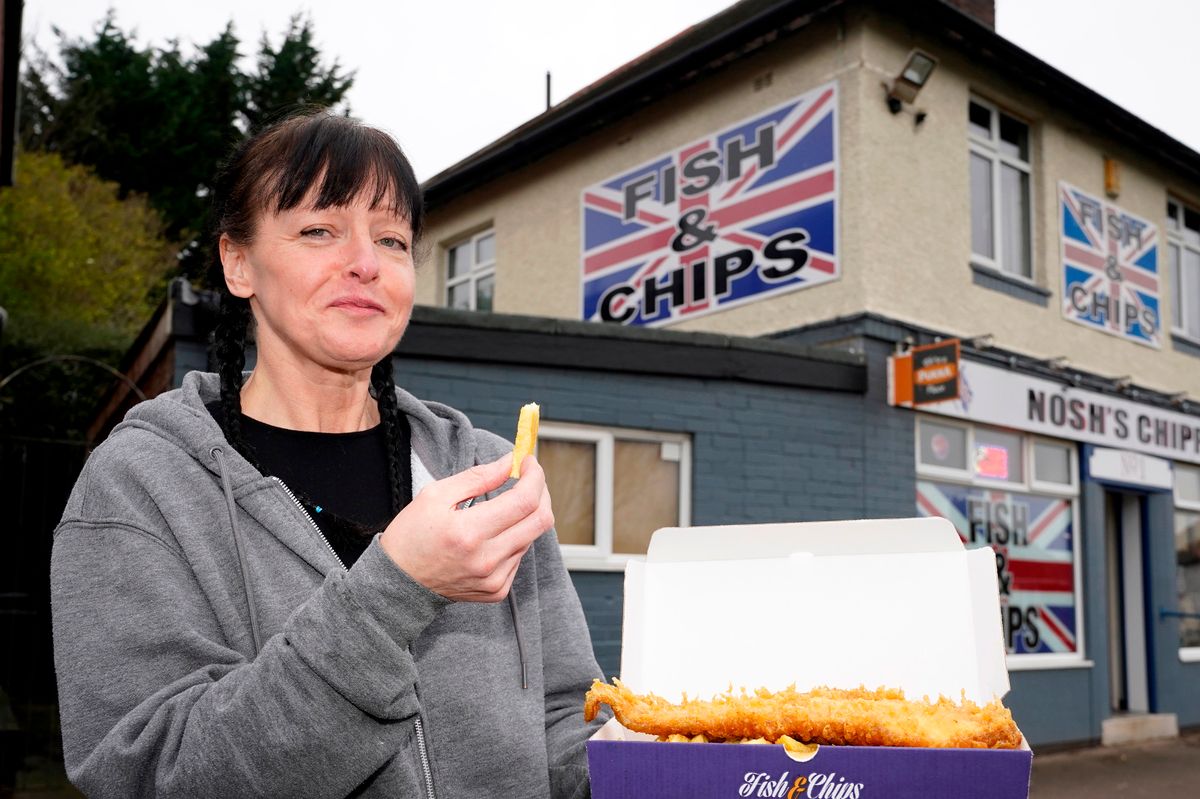 Sam stands outside the house in front of the chippie in Sheffield holding a box of chips with one in her hand.