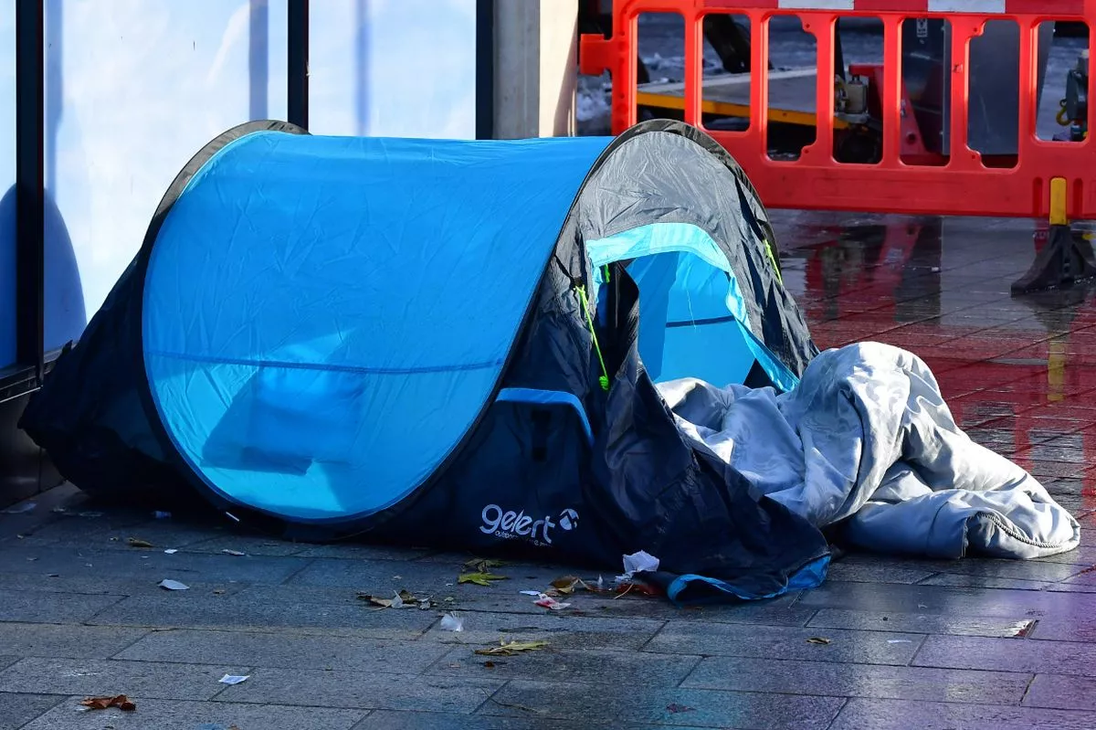 A homeless person's tent in Liverpool City Centre. The blue tent has it's front open and a sleeping bag is spilling out onto the pavement. The floor is wet as it has been raining and there are leaves and other rubbish blown up against the side of the tent.