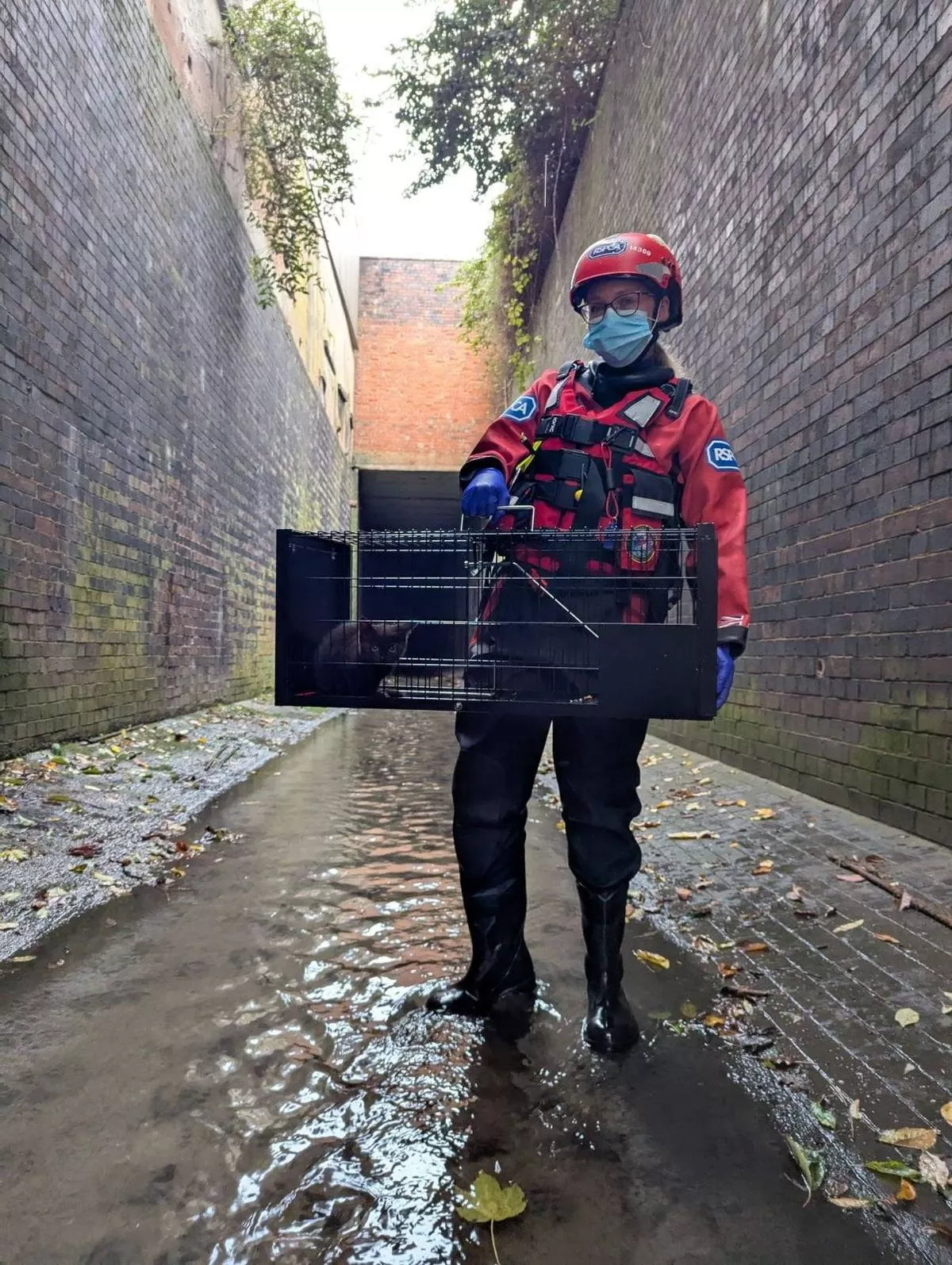 An individual dressed in a red jacket, helmet, and mask is standing in a wet, narrow alleyway between two brick walls.