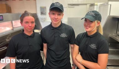 Two young women and one young man (centre) in their chip shop uniforms of a black polo shirt and trousers and a skip cap smile for the camera. They are standing beside the fat fryers and food prep area, all in stainless steel.