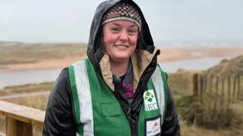 Claire Stainfield smiling at the camera with Newburgh beach in Aberdeenshire behind he, she is wearing a jacket and a wool knitted hat.