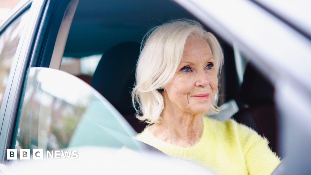 An older woman driving a car. She has blonde hair and is wearing a light yellow jumper.