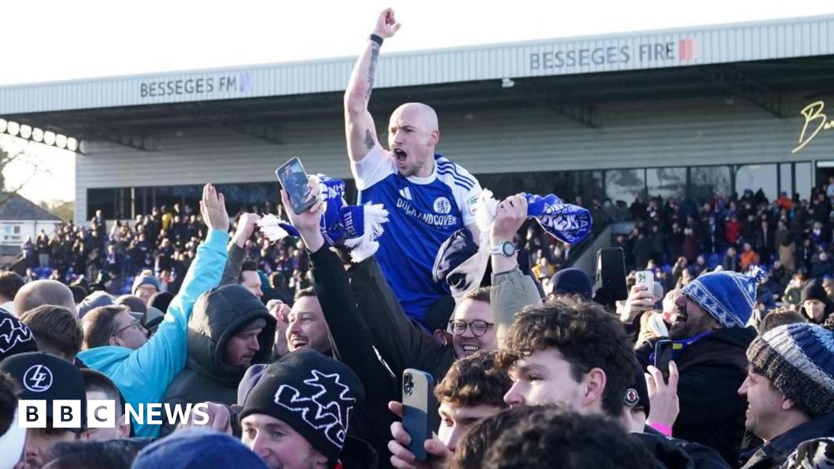 Macclesfield's Josh Kay raises his arm in the air celebrating while surrounded by fans on the pitch. His mouth is open and he looks to be cheering. He appears to be on the shoulders of a team-mate and raised above the surrounding fans.