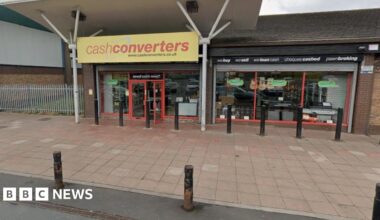 The front of a Cash Converters store. The sign indicating the shop is yellow, with red writing. A large pavement with bollards is visible in front of the store.