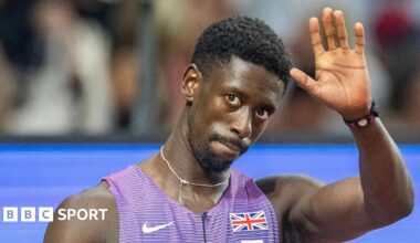 Former Great Britain sprinter Reece Prescod waves with his left hand as he wears purple and white kit with a union jack badge and Nike swoosh logo on the front