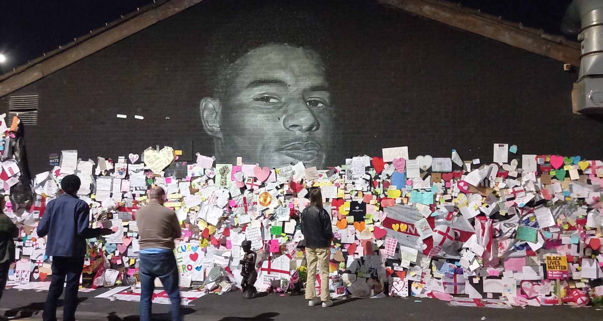 A black wall with a mural of the face of footballer Marcus Rashford on it, the wall is covered in thousands of pieces of paper in all colours, shapes and sizes