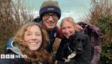 The family huddle together and smile at the camera. They are in outdoor walking gear and the sea is in the background. Tara, the mother, has the family's black and white dog on her lap.