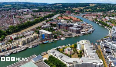 An aerial image capturing Bristol’s Harbourside area with the River Avon winding through the city. There are colourful residential houses and modern waterfront apartments, alongside a variety of boats docked along the quays. In the distance there are fields, hills and woodland. It looks like a warm and sunny day and the water is a deep green.