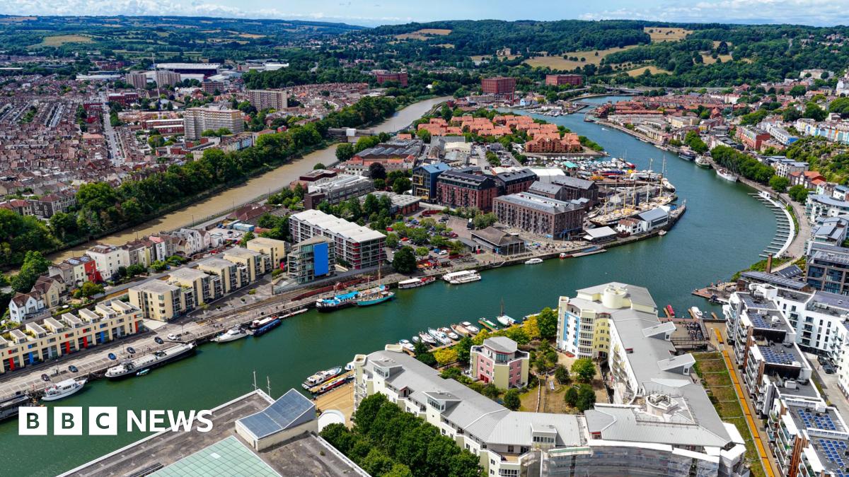 An aerial image capturing Bristol’s Harbourside area with the River Avon winding through the city. There are colourful residential houses and modern waterfront apartments, alongside a variety of boats docked along the quays. In the distance there are fields, hills and woodland. It looks like a warm and sunny day and the water is a deep green.