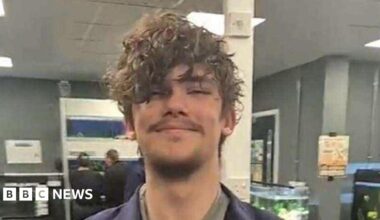 A young man who has brown curly hair and some short facial hair is pictured smiling in an office-type room that appears to have several large tropical fish tanks in.