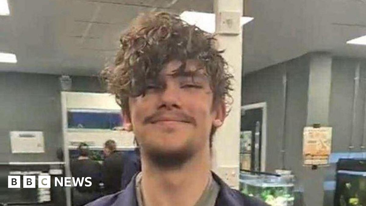 A young man who has brown curly hair and some short facial hair is pictured smiling in an office-type room that appears to have several large tropical fish tanks in.