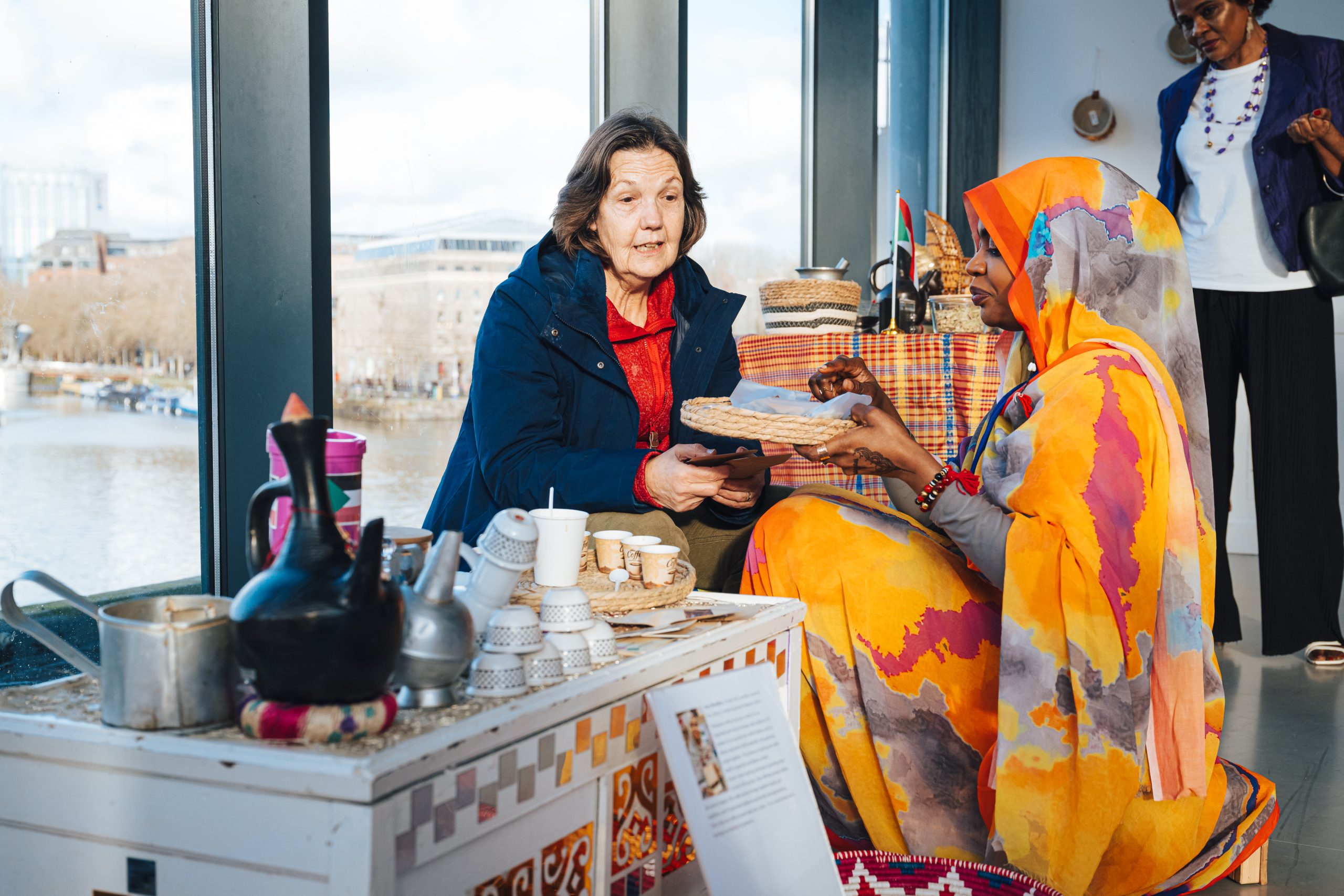 Two women sit in front of a large window next to a coffee stand