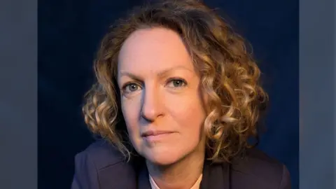 BBC A professional headshot of Victoria Woodall with chin length golden curly hair wearing a navy suit against a navy backdrop