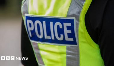 The back of a police officer who is wearing a hi-vis police vest over a black top.