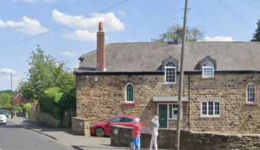 Two people stand at the corner of two roads on a sunny day. There are large stone-coloured houses and a red car parked on the drive.