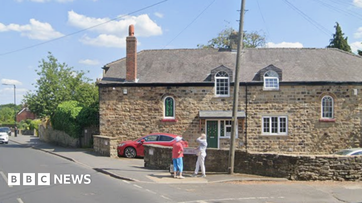 Two people stand at the corner of two roads on a sunny day. There are large stone-coloured houses and a red car parked on the drive.