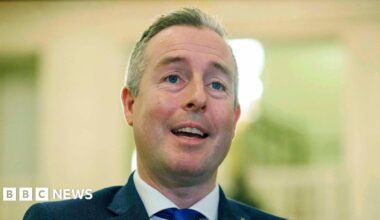 Paul Givan is wearing a navy blazer, white shirt and blue tie. He has short grey hair. The background is blurred.