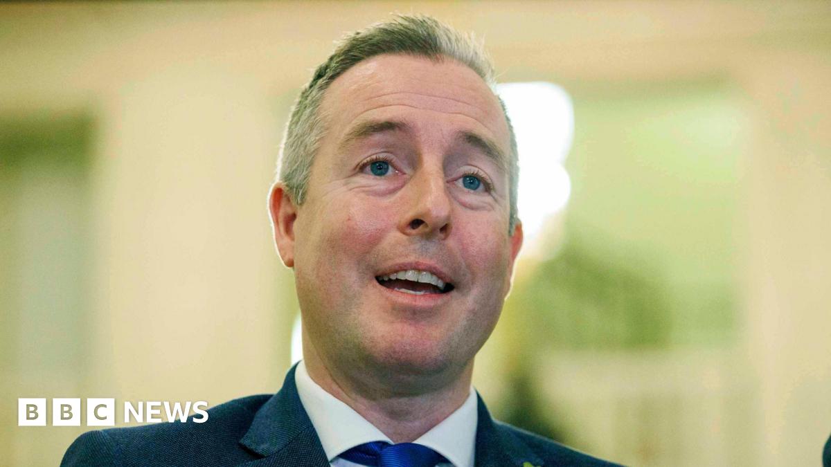 Paul Givan is wearing a navy blazer, white shirt and blue tie. He has short grey hair. The background is blurred.