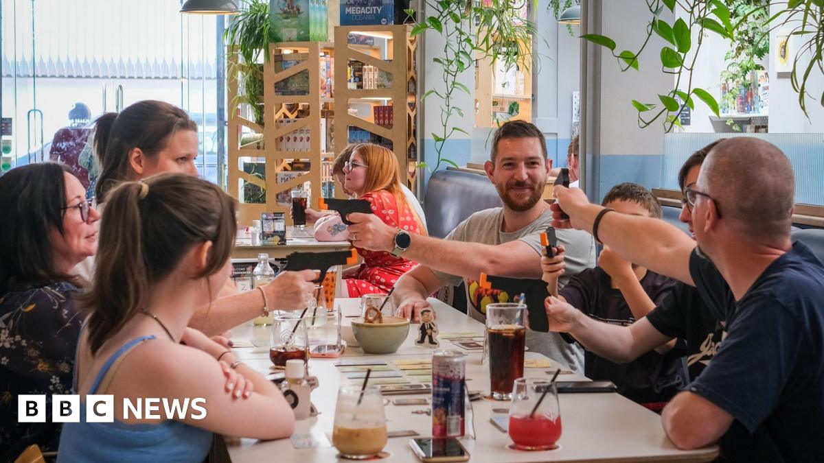A general view of the cafe. In the foreground are six people sitting around a table. A few of them are pointing plastic guns at each other. There are cards set out on the table. Everyone looks amused.