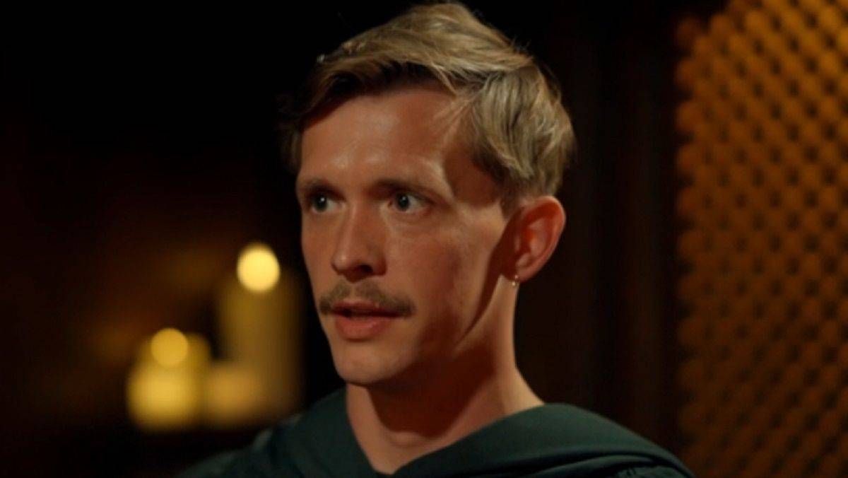 An individual with a mustache and short, light-colored hair, dressed in a dark-colored garment, is standing in a dimly lit environment. The background features subtle lighting, creating a soft contrast against the subject.