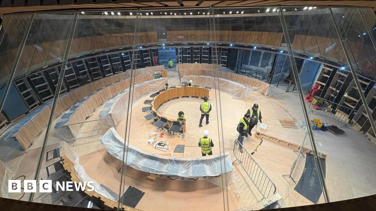 The image shows construction workers at work in the Senedd debating chamber. The politicians' desks are covered in polythene.