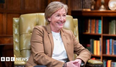 A close up shot of a middle-aged blonde woman sat in a leather chair in a room with a bookcase behind her smiling - Amanda Collier on The Traitors