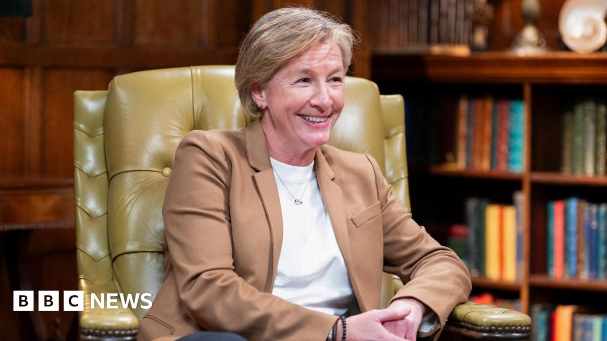 A close up shot of a middle-aged blonde woman sat in a leather chair in a room with a bookcase behind her smiling - Amanda Collier on The Traitors