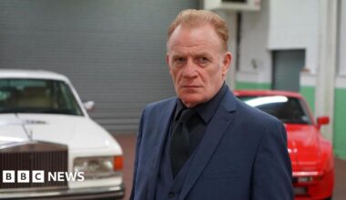 Mark Lewis Jones, wearing a blue two piece suit with a dark shirt. He has light brown/ginger hair. Pictured behind him are two cars; one white and one red.