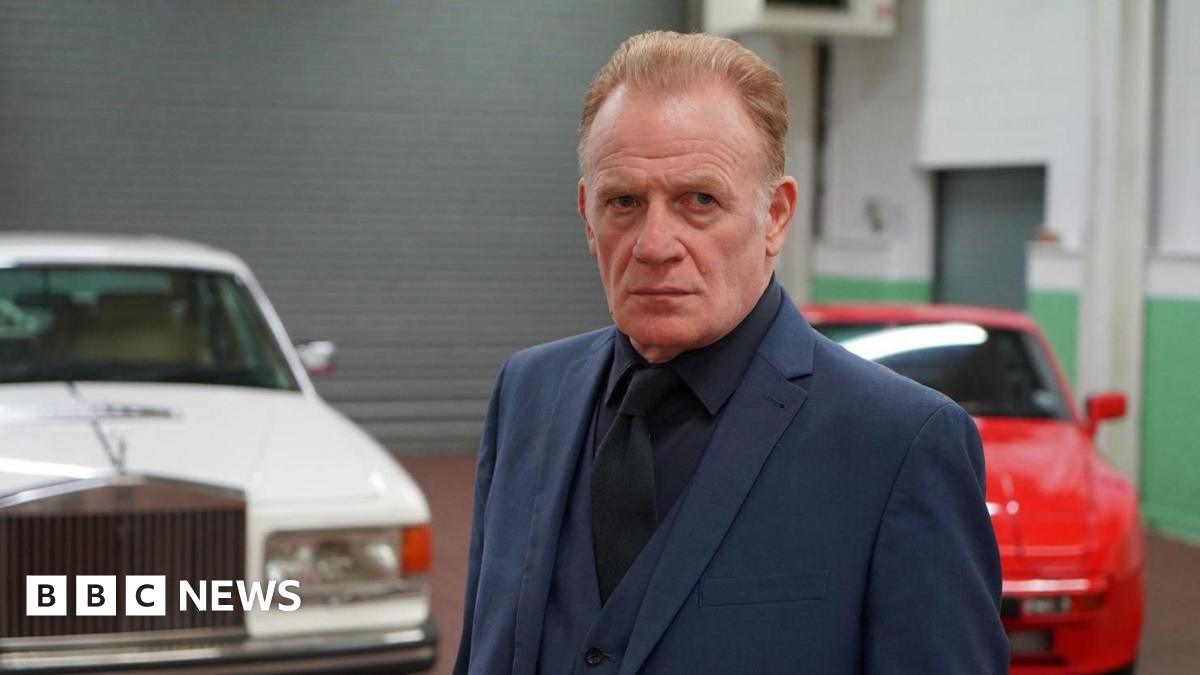 Mark Lewis Jones, wearing a blue two piece suit with a dark shirt. He has light brown/ginger hair. Pictured behind him are two cars; one white and one red.