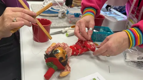 BBC News A wooden puppet wearing a red costume and hat lies on the table. It has green eyes and brown hair. A lady wearing pink is examining its strings while another hand holds up its wooden top.