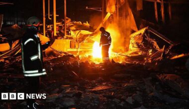 Emergency workers stand in front of a blaze at night. reflective strips on their coats stand out in the shadows and light from the fire.
